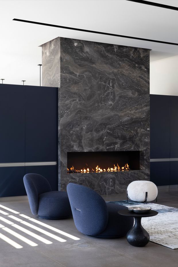A sophisticated modern living room centred on a dramatic full-height dark veined stone fireplace with a long linear gas fire. Deep navy built-in wall panelling with slim horizontal trim, a white ceiling with recessed linear details, sculptural navy lounge chairs, a round black side table, and a pale textured rug create a refined, high-contrast interior.