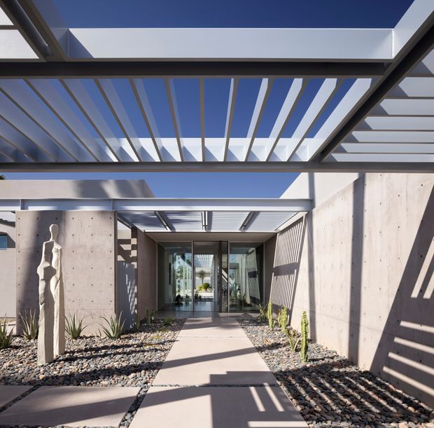 A modern architectural entry courtyard viewed from outdoors, with a concrete pathway leading to glazed doors beneath layered steel or aluminium pergolas. The composition features strong geometric lines, deep overhangs, patterned shadows, exposed concrete walls, gravel landscaping, and sparse drought-tolerant planting. A sculptural stone or concrete artwork stands beside the entrance under a clear blue sky.