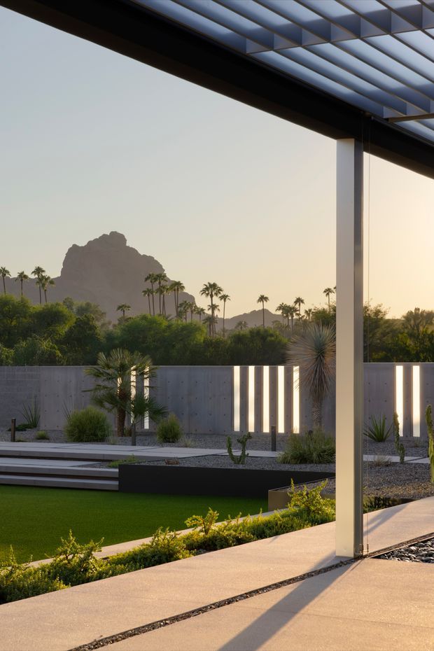 A contemporary covered outdoor terrace or patio overlooking a landscaped desert garden. The scene features a slim dark roof structure with translucent or glazed paneling, a tall square support column, pale concrete paving, linear gravel drainage strips, synthetic or manicured lawn, sculptural desert planting, and a concrete privacy wall with illuminated vertical slots. Palm trees and a distinctive rocky mountain formation are visible beyond the property in warm late-afternoon light.