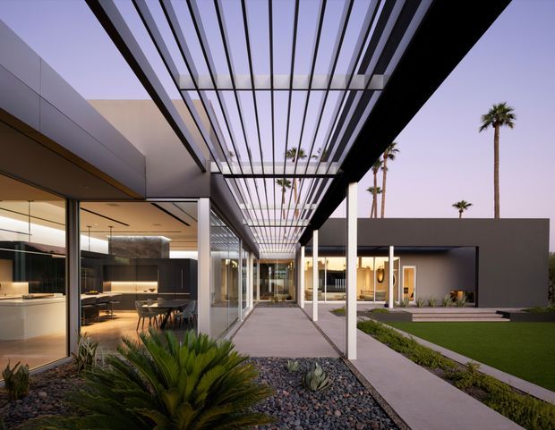 A contemporary single-level residence arranged around a landscaped courtyard. The image looks along a paved outdoor walkway beneath a dramatic black steel pergola with closely spaced linear slats. Full-height glazing reveals a warm, sophisticated kitchen and dining area on the left, while a minimalist cubic wing and lawn sit to the right. Palm trees, gravel landscaping, and architectural planting create a refined resort-like outdoor setting.