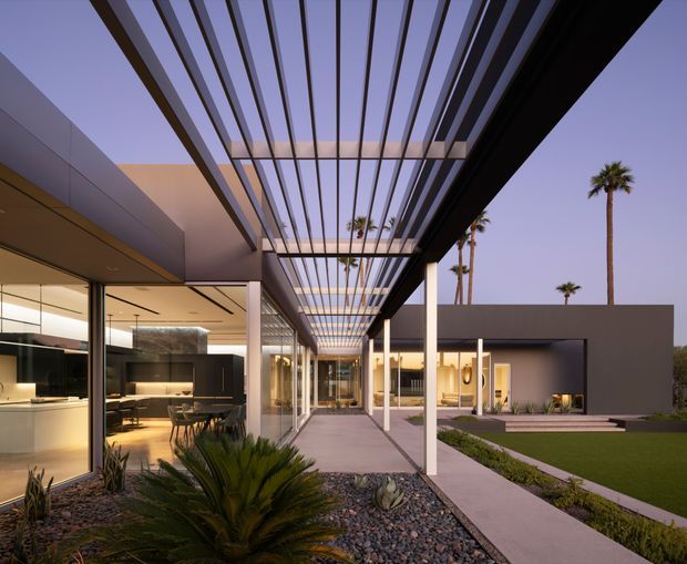 A sophisticated modern single-storey residence viewed from a landscaped exterior walkway and covered courtyard. The composition is dominated by a long pergola with closely spaced dark metal slats, creating a strong linear canopy over a paved path. Floor-to-ceiling glazing reveals a warm, refined interior with a kitchen and dining area, while white structural columns, smooth plastered walls, gravel landscaping, and clipped lawn surround the home. Palm trees and a dusky sky are visible beyond the property.