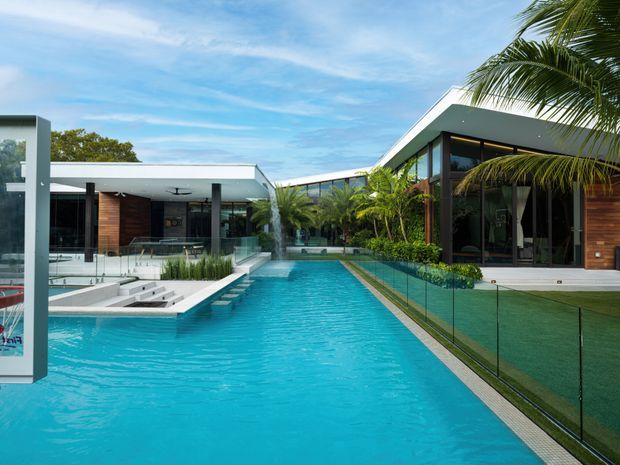 A contemporary single-storey residence arranged around a long turquoise swimming pool. The view looks across the pool toward low flat-roofed wings with extensive dark-framed glazing, timber-clad wall sections, white roof planes, glass balustrades, and lush tropical planting. The poolside setting is bright and resort-like, with a strong indoor-outdoor connection.