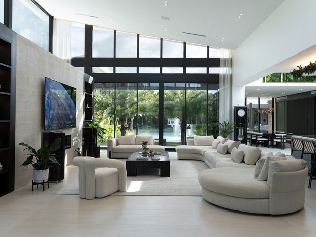 Bright, spacious contemporary living room with a double-height glazed facade overlooking greenery and a pool. The room features a large curved cream sectional sofa, matching sculptural lounge chairs, a low black coffee table, a pale area rug, dark built-in shelving, a textured light stone or concrete feature wall with wall-mounted artwork, and an open dining area beyond.