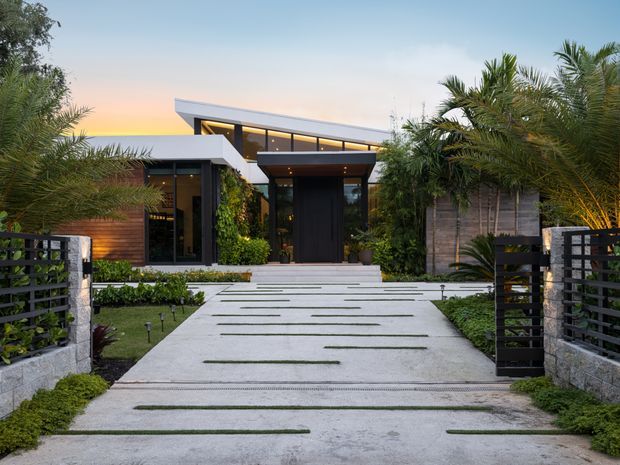 A contemporary single-storey house viewed from the front driveway at dusk. The home features strong horizontal roof planes, extensive dark-framed glazing, timber wall cladding, a recessed entrance, lush tropical landscaping, and a broad pale concrete driveway with narrow linear planting joints. Stone and dark horizontal-slat boundary walls frame the approach.