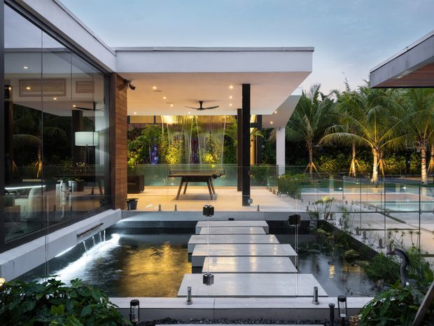 A sophisticated contemporary residence opening onto a landscaped courtyard and pool area. The view is framed by expansive floor-to-ceiling glazing, white flat-roofed slabs, dark structural columns, timber wall cladding, and a glass balustrade. A shallow reflective water feature with stepping slabs leads toward the covered outdoor entertaining space, with dense tropical planting surrounding the swimming pool.
