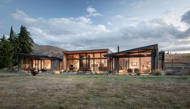 A single-storey contemporary home set in an open rural or alpine landscape, photographed from the front at dusk. The building is composed of several low, connected flat-roofed volumes with extensive floor-to-ceiling glazing, dark metal framing, warm timber-toned interiors, and sheltered outdoor areas. A mountain ridge and conifer trees form the background, while the broad grass-covered foreground emphasizes the home's relationship to the landscape.