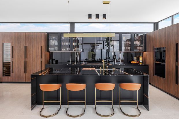 A sophisticated contemporary kitchen with a large black island and four orange bar stools. Dark timber cabinetry spans both side walls, while black stone surfaces, glass-fronted upper cabinetry, integrated appliances, and reflective splashback glazing create a sleek, high-contrast interior. A high ceiling and clerestory glazing bring in abundant natural light.