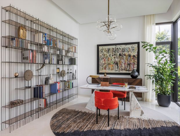 A bright, refined home office with a sculptural open metal bookcase spanning the left wall, a marble-topped desk, mid-century-style timber credenza, red upholstered chairs, large abstract artwork, a statement glass-and-metal pendant, and a tall indoor plant beside full-height corner glazing. The room has a contemporary shell with an eclectic mid-century and luxe character.