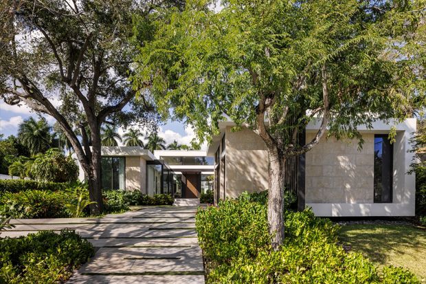 A low-profile modern residence set within mature tropical landscaping. The facade combines pale stone or concrete walls, dark-framed floor-to-ceiling glazing, and crisp flat-roofed forms. A paved walkway leads through hedges and shaded trees toward a recessed entry, creating a strong indoor-outdoor connection.