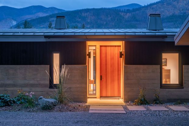 Exterior dusk view of a low-slung contemporary home with dark vertical cladding, a warm timber entry door, concrete privacy walls, roof-mounted skylight or vent structures, and illuminated windows. A gravel and planted entry landscape leads toward the house, with wooded mountains visible in the background.