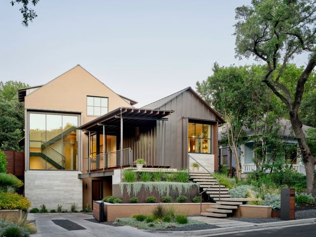 Exterior view of a contemporary multi-level residence composed of two connected gabled volumes. The home features warm beige rendered walls, dark vertical timber cladding, extensive glazed openings, a covered balcony or entry porch, and prominent exterior stairs leading through layered landscaped garden beds. Mature trees and low-water planting surround the house.