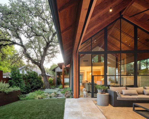 A contemporary indoor-outdoor living space opening onto a landscaped courtyard. The image features a large glazed gable end with dark metal framing, warm timber-lined soffits and ceiling, exposed structural beams, and a covered stone-paved terrace furnished with an outdoor sectional sofa and coffee table. Mature trees and planted garden beds surround the dwelling, while the interior living area is visible through the glazing.