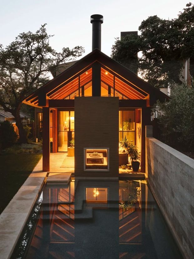 A dramatic dusk view of a compact contemporary house with a steep gable roof and deep dark eaves. Warm linear lighting illuminates the exposed timber roof structure and glazed living spaces. A tall central dark masonry chimney with an integrated fireplace dominates the foreground, reflected in a long narrow reflecting pool bordered by concrete paving and a rendered boundary wall. Mature trees surround the dwelling.