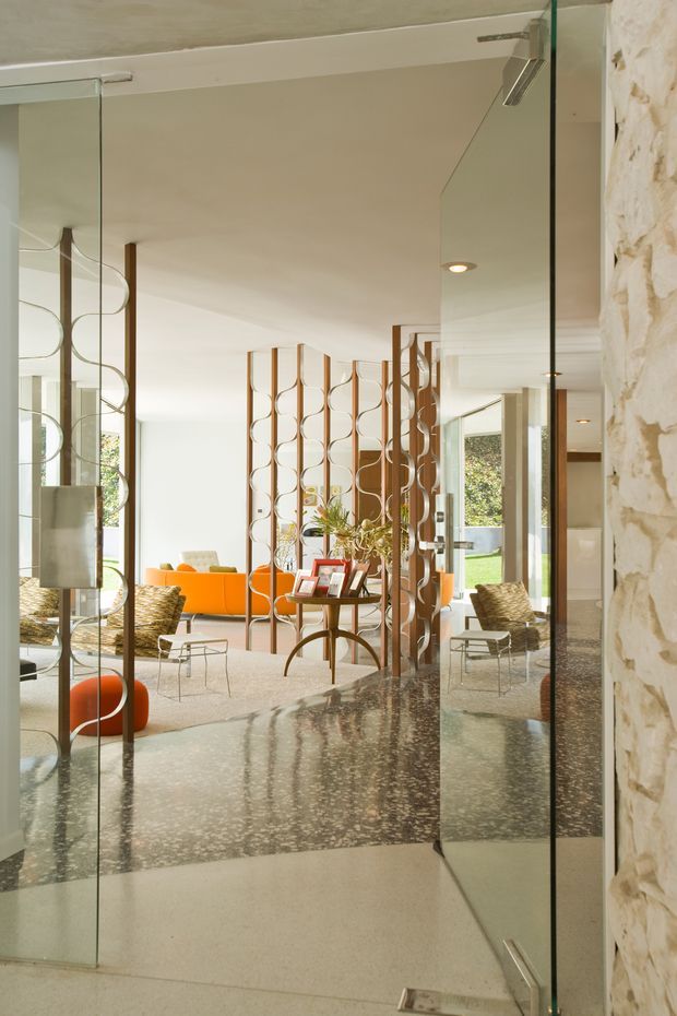 View through frameless glass doors into a bright, refined mid-century-inspired living room. The space features a terrazzo-like polished floor, cream walls and ceiling, a sculptural open wood room divider with repeating circular motifs, orange upholstered seating and a central timber table. A textured pale stone wall frames the right side, while garden greenery is visible beyond the glazing.