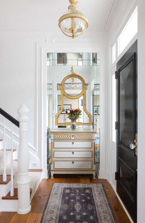 Bright, refined entry hallway with white paneled walls, a staircase and balustrade on the left, a dark paneled door on the right, and a central decorative console with drawers beneath an ornate gilded mirror. A patterned runner extends along the polished timber floor, while a traditional gold pendant light hangs overhead. Reflections in the mirror reveal additional interior detailing and glazing.
