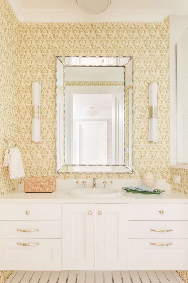 A bright, elegant bathroom vanity with a wide white built-in cabinet, integrated basin, and polished metal fixtures. Pale patterned wallpaper covers the walls, flanked by two vertical wall sconces. A large framed mirror reflects a white paneled doorway and ceiling light. The room has traditional detailing with warm gold hardware and a light, refined palette.