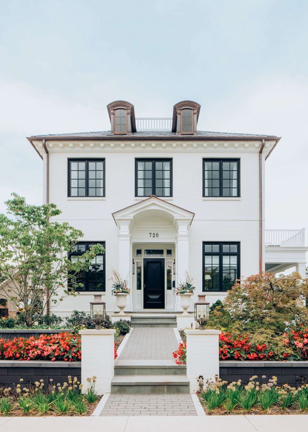 A symmetrical two-storey white masonry residence viewed from the front, with black multi-pane windows, a prominent classical portico entrance, decorative cornice detailing, dormer windows and a small rooftop balcony. A formal landscaped garden with paved paths, raised planters and colourful flowering beds leads to the entry.
