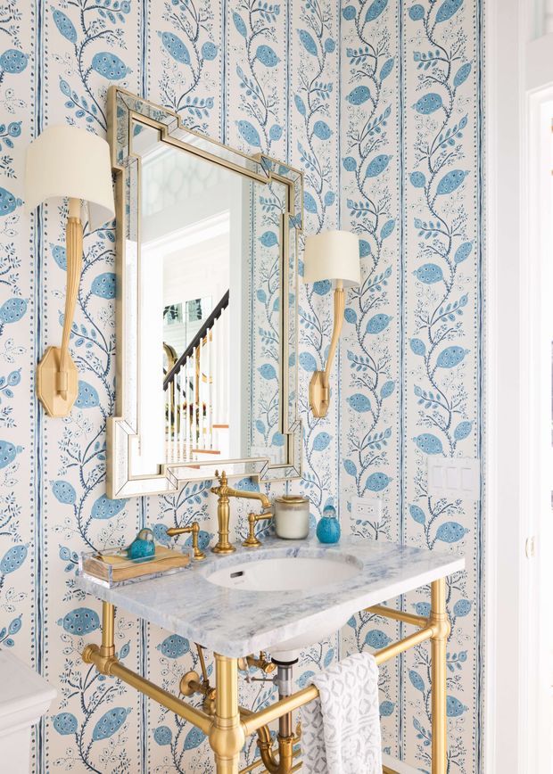 Bright, compact bathroom vanity with a white marble or stone benchtop, exposed brass plumbing, brass tapware, and a large geometric framed mirror. The walls are covered in a blue-and-white botanical patterned wallpaper, complemented by matching blue accessories and brass wall sconces with cream shades. A toilet is partially visible at left, and a staircase is reflected in the mirror.