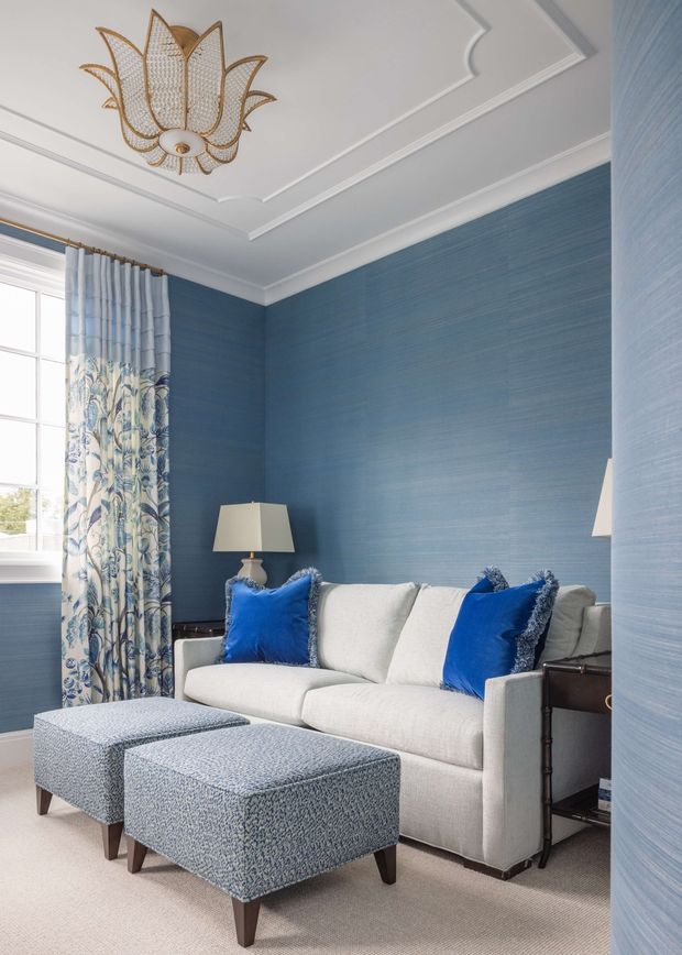 A refined, compact living room with deep blue textured wallcoverings, a pale upholstered sofa, vivid blue accent cushions, patterned blue-and-white ottomans, and coordinated botanical curtains. The room features a decorative gold-toned floral pendant, layered white ceiling moulding, classic table lamps, and soft neutral carpeting. Natural light enters through a large window, creating an elegant and inviting atmosphere.