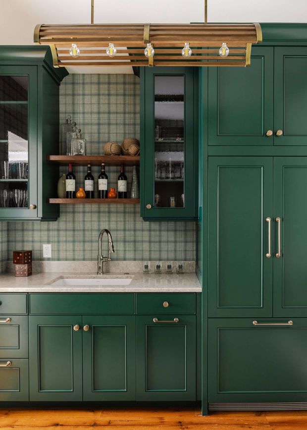 A richly detailed traditional-style kitchen with deep forest-green shaker cabinetry, brass knobs and pulls, a pale stone benchtop, and a green-and-beige checkered tile backsplash. Open shelving displays bottles and glassware, while a tall integrated cabinet occupies the right side. A timber-and-brass linear pendant light hangs above the work area, and warm timber flooring grounds the space.