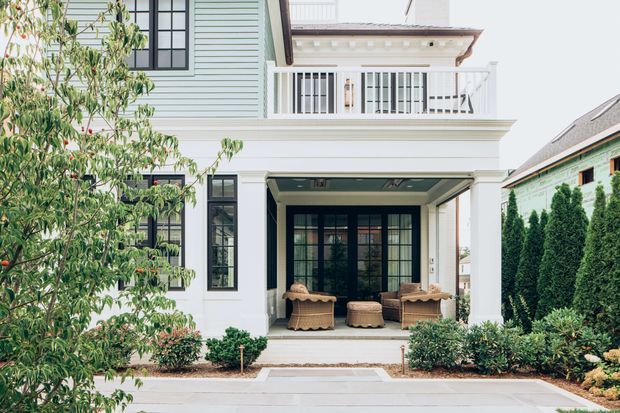 Front exterior view of a refined two-storey home with pale horizontal weatherboard cladding, white classical trim, black-framed divided-light windows and doors, and a covered ground-floor porch furnished with wicker seating. The upper level has a small balustraded balcony, while clipped hedges, shrubs, and a paved front path frame the facade.