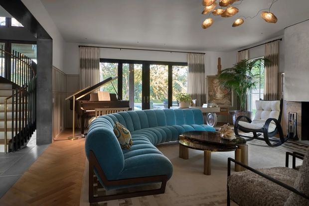 A sophisticated living room with a large curved teal upholstered sectional sofa, a dark wood and glass coffee table, a white lounge chair, and a sculptural black rocking chair. The room features full-height glazed doors and windows with curtains, wood wall panelling, a piano near the glazing, a textured stone fireplace wall, and a striking organic pendant light. A stairway with dark balustrading is visible at left, while herringbone timber flooring transitions to pale carpet in the seating area.