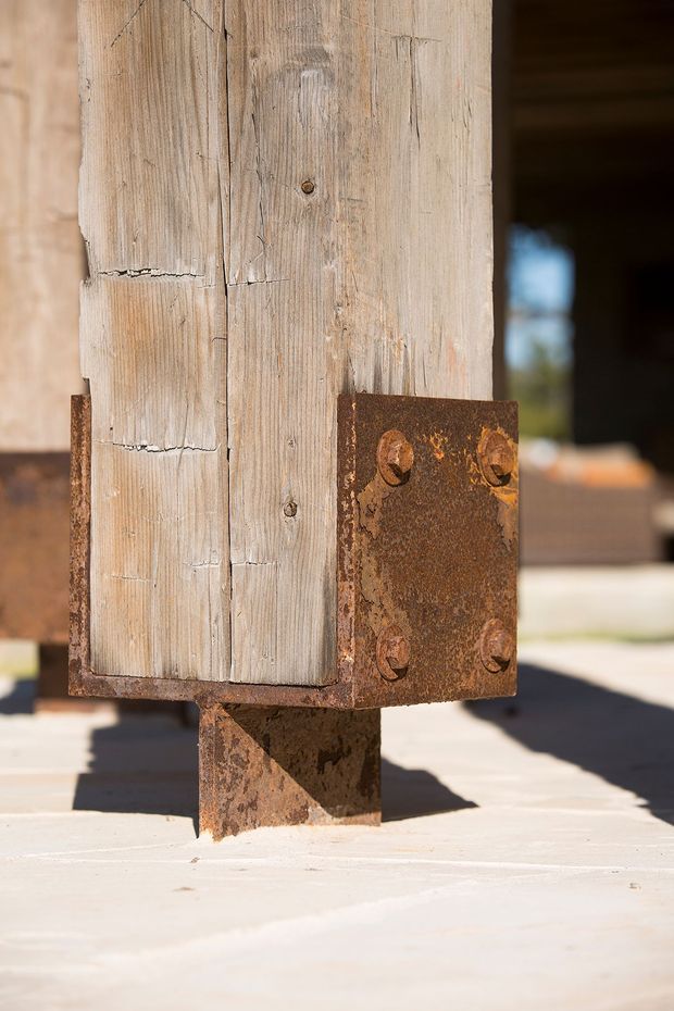 Close-up architectural detail of a weathered timber post reinforced with a rusted steel bracket and bolted connection. The timber and corroded metal show raw, aged textures, with a blurred building opening and sunlit paved surface in the background.