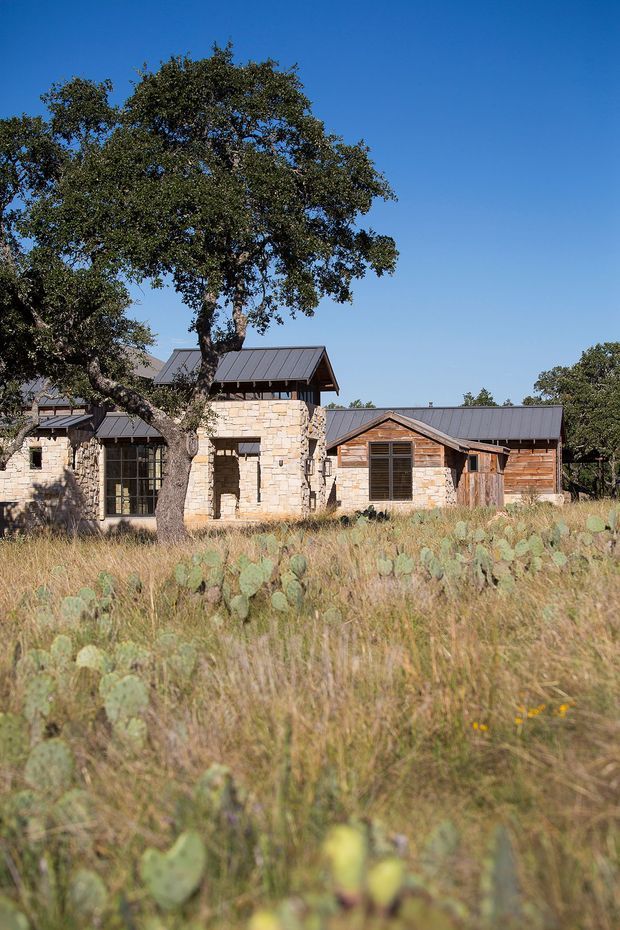 Exterior view of a low-rise rustic residence set in a dry natural landscape, with pale stone walls, timber-clad gabled sections, dark standing-seam metal roofs, and large glazed openings. Mature trees and prickly-pear vegetation surround the building under a clear blue sky.
