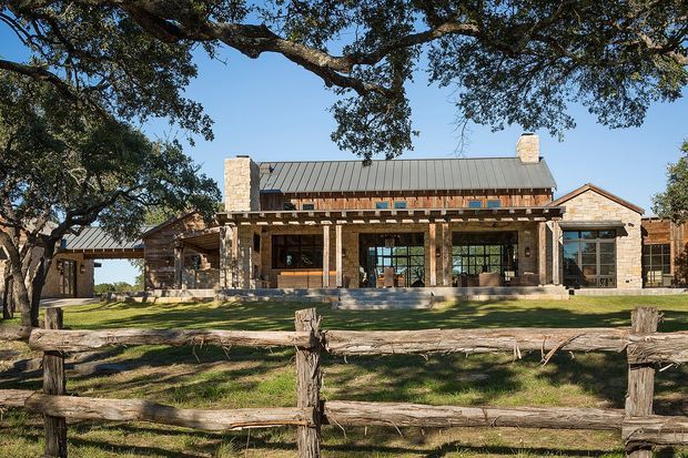 Exterior view of a broad, single-storey stone house with a long covered veranda, timber posts and beams, dark standing-seam metal gable roof, masonry chimneys, and large multi-pane windows. A rustic split-rail timber fence and mature oak trees frame the landscaped foreground. The building has a substantial ranch or farmhouse-like form with natural stone and timber detailing.