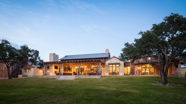 A wide exterior view of a single-storey residential home at dusk, set behind a broad lawn and mature trees. The house features extensive warm-lit glazing, natural stone masonry walls and chimneys, timber detailing, and a low-pitched standing-seam metal roof. A central glazed living area is flanked by stone and timber-clad wings, creating a rustic yet refined ranch-style composition.