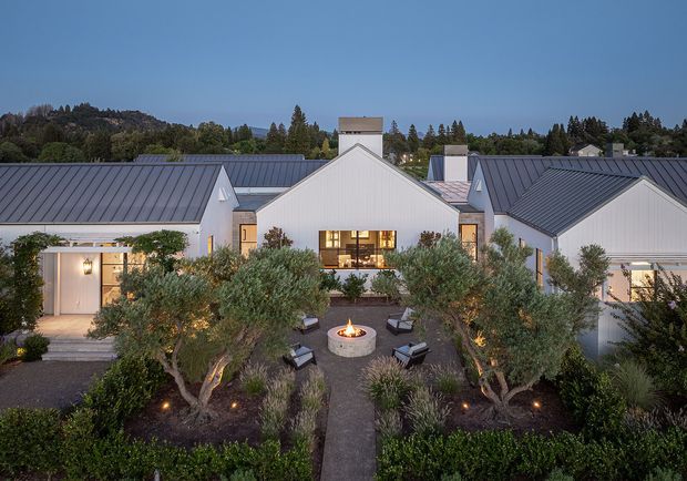A dusk aerial view of a low-rise white residential complex arranged around a landscaped central courtyard. The buildings feature crisp vertical cladding, dark standing-seam pitched roofs, gabled forms, large illuminated windows, and a central chimney-like element. The courtyard includes olive trees, clipped hedges, gravel planting beds, a straight paved path, lounge chairs, and a circular fire pit, with wooded hills and neighboring homes in the background.