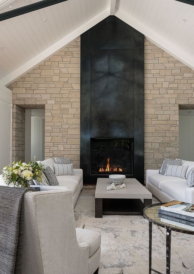 A refined living room with a dramatic full-height black metal fireplace surround set into a pale stone-clad gable wall. White-painted vaulted ceilings and exposed-looking panel joints frame the symmetrical composition, with two door openings on either side of the fireplace. The room contains soft white sofas and armchairs, a pale rectangular coffee table, a patterned rug, and a glass-topped side table with books. The overall scheme is light, neutral, and elegant with strong contrast from the fireplace.