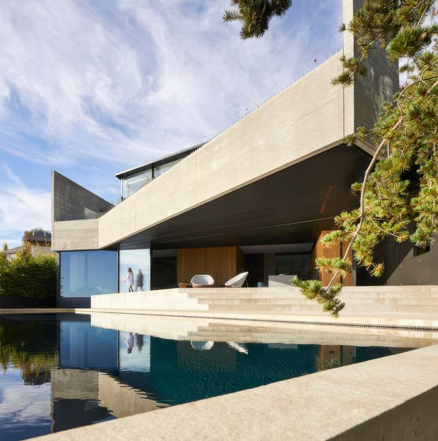 A contemporary multi-level residence viewed from beside a reflective swimming pool. The house features strong cantilevered horizontal concrete forms, extensive glazing, a deep shaded outdoor living area, warm timber wall cladding, and broad pale stone or concrete terraces. Pine-like landscaping frames the right side of the image, while the pool mirrors the architecture and sky.