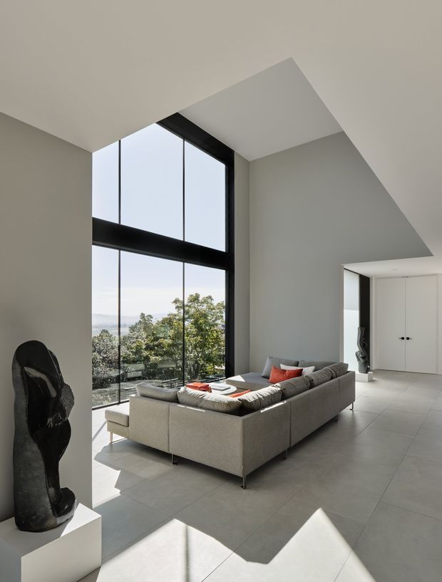 A spacious contemporary living room with a large grey sectional sofa, sculptural black artwork, and expansive floor-to-ceiling glazing overlooking a leafy landscape. The room has a dramatic angular ceiling, pale tiled flooring, white walls and built-in white cabinetry, with strong natural light creating geometric shadows.