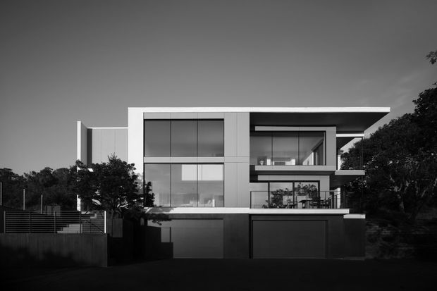 Black-and-white exterior photograph of a large contemporary multi-storey residence. The house has a strongly rectilinear composition, flat roof planes with broad overhangs, extensive floor-to-ceiling glazing, pale wall cladding, dark garage doors, and projecting upper-level volumes. Dense trees and boundary fencing frame the property.