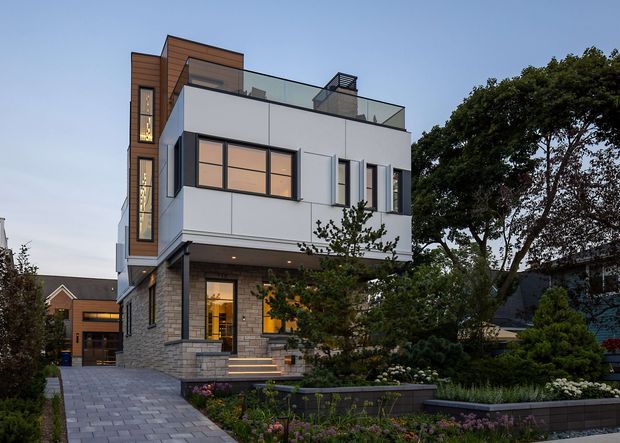 Exterior view of a contemporary multi-storey urban residence with a predominantly white rendered facade, projecting upper level, rooftop terrace with glass balustrade, warm timber-clad vertical feature volume, and dark-framed windows. The house is set along a paved driveway and landscaped front garden, with neighbouring homes visible around it.