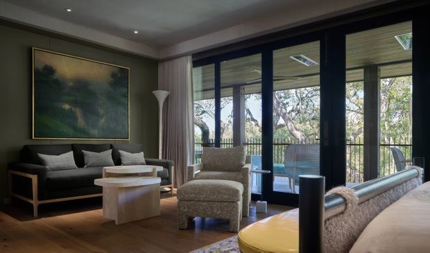 A refined living room with dark green-grey walls, a black sofa, sculptural light wood coffee tables, a textured lounge chair and ottoman, and a large framed abstract artwork. Full-height dark-framed glazed doors open to a balcony or terrace with views of surrounding trees. Warm timber flooring, layered neutral upholstery, recessed ceiling lights, and soft curtains create a sophisticated, calm atmosphere.