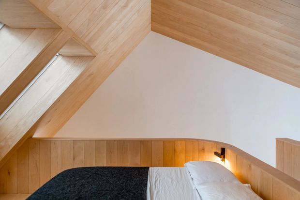 A compact attic bedroom beneath a sloping roof, featuring warm light timber wall panelling, exposed timber ceiling and roof structure, white plasterboard, and a partially visible bed with white bedding and a dark textured throw. Integrated wall lighting creates a warm, intimate atmosphere.