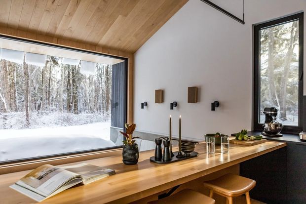 A warm, contemporary kitchen with a long light-wood island or benchtop, black lower cabinetry, and a large black-framed corner window overlooking a snowy forest. The ceiling is clad in matching timber boards, while minimalist black wall-mounted fittings, pendant-free candleholders, stools, and small countertop appliances create a refined cabin-like interior.