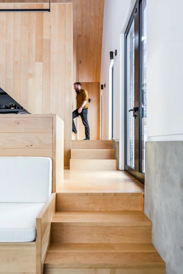 Interior view of a compact, bright modern home with a light timber staircase and integrated timber joinery. The staircase rises beside full-height dark-framed glazing, while a built-in white upholstered bench and partial kitchen cabinetry are visible to the left. Smooth white plaster and a grey concrete-like wall contrast with the pale wood surfaces.