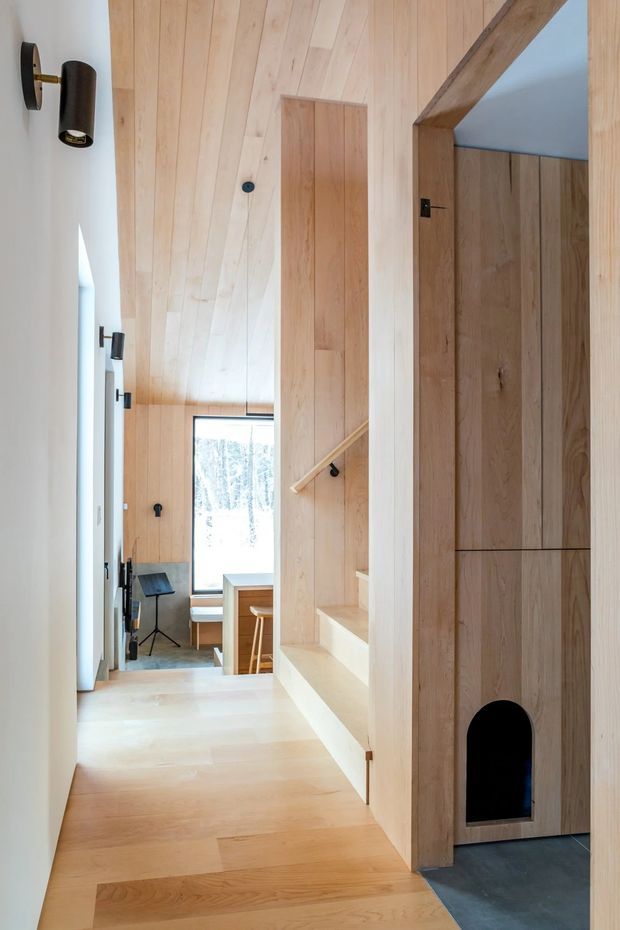 A narrow, light-filled residential hallway clad extensively in pale timber boards, with matching timber flooring and integrated cabinetry. A compact staircase with a simple timber handrail rises beside the passage, while a tall timber storage unit includes a small arched opening at its base. The corridor opens toward a bright workspace or living area with a window, desk, and stool. Minimal black wall sconces and hardware provide subtle contrast against the warm wood and white walls.
