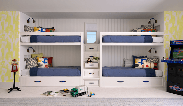 A bright, built-in bunk room with two symmetrical white bunk-bed units, each containing upper and lower bunks with blue bedding, patterned and colourful cushions, integrated reading lights, and drawers beneath. A central column of built-in drawers separates the bunks, while a patterned yellow-and-white wall finish, scattered children's toys, a toy vending machine, and a pinball arcade cabinet create a playful children's retreat. The room has a low, standard ceiling and a light neutral carpet.
