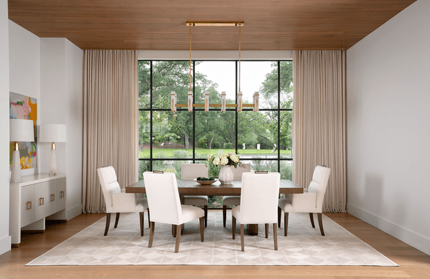 A bright, refined dining room with a long timber dining table, six upholstered dining chairs, a geometric area rug, and a linear pendant chandelier. Full-height curtains frame large black-framed windows overlooking a leafy garden. The room features a warm timber ceiling and floor, white walls, and a low built-in sideboard with decorative objects.