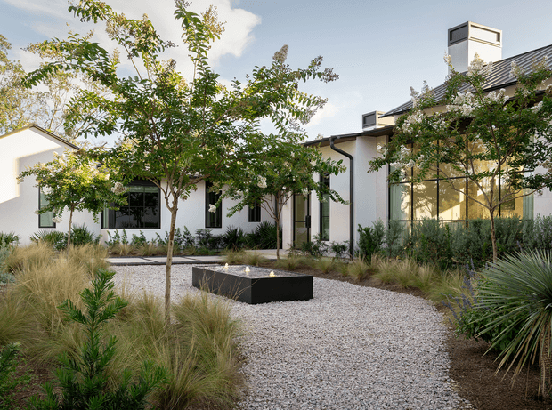 Exterior view of a contemporary single-storey residence framed by a landscaped courtyard garden. White rendered walls, dark-framed glazing, a dark pitched roof, and prominent rooftop ventilation or chimney structures surround a pale gravel pathway and a low rectangular black water feature or fire feature. The planting is drought-tolerant and architectural, with ornamental grasses, agaves, and small trees.
