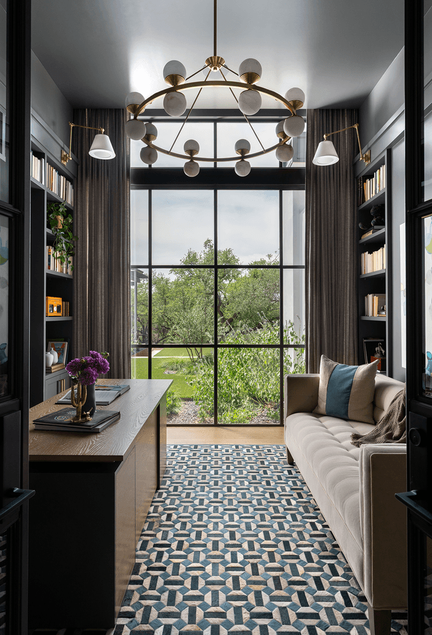 A narrow, sophisticated home office or study viewed through dark-framed glass doors. The room has built-in black shelving on both sides, a substantial wood-topped desk, a cream upholstered sofa, dark floor-to-ceiling curtains, and a large black-framed window overlooking greenery. A sculptural brass-and-globe pendant light and matching wall lamps provide a refined focal point, while a geometric blue, cream, and grey patterned rug adds visual interest.