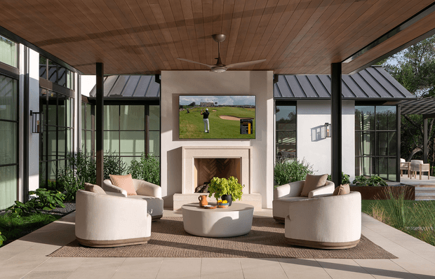 A refined indoor-outdoor living room with expansive black-framed glazing, a warm timber-lined ceiling, central fireplace and television wall, and curved cream lounge chairs arranged around a sculptural coffee table. Planting beds flank the seating area, while glazed extensions and a covered outdoor area are visible beyond.