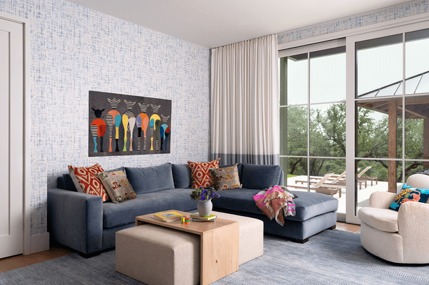 A bright contemporary living room with a large charcoal-blue L-shaped sectional sofa, patterned accent cushions, a pale wood square coffee table, and a matching upholstered ottoman. The room features blue-and-white textured wallpaper, full-height cream curtains, white trim, expansive black-framed glazed doors or windows, and a large colorful figurative artwork. A patio and greenery are visible beyond the glazing.