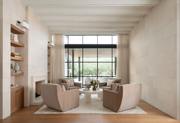 A refined, light-filled living room with a symmetrical seating arrangement around a round coffee table. Four upholstered lounge chairs sit on a large pale rug over warm timber flooring, with built-in timber shelving and cabinetry flanking a fireplace on the left. Full-height curtains frame expansive black-framed glazing, while the ceiling features exposed painted beams and the walls are clad in large pale stone or tile panels.