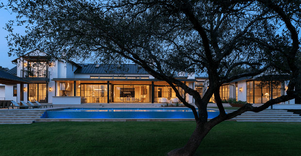 A wide dusk exterior view of a large contemporary residence arranged around a long illuminated swimming pool. The home has a low-pitched dark metal roof, extensive floor-to-ceiling glazing, white rendered walls, warm interior lighting, broad stepped terraces, and a mature spreading tree dominating the foreground. The manicured lawn and pool create a refined landscaped courtyard-like setting.