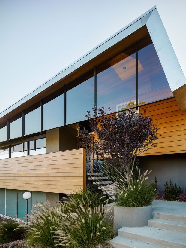 Low-angle exterior view of a modern multi-level residence with extensive floor-to-ceiling glazing, dark slender window frames, warm horizontal timber cladding and soffits, and a crisp angular roofline. A prominent exterior stair with metal railings rises through landscaped ornamental grasses and raised concrete planters, while a timber privacy screen partially encloses the lower level.